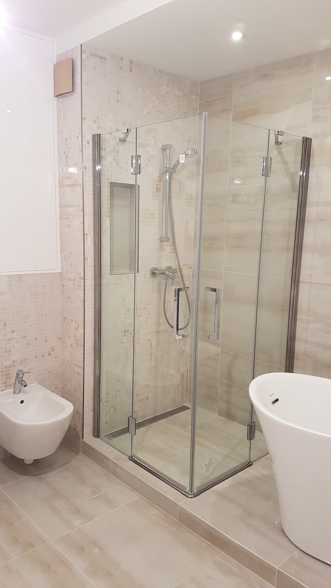 Nowoczesna kabina prysznicowa z transparentnego szkła, chromowanymi zawiasami i odpływem liniowym, obok białe bidet i wanna wolnostojąca, całość w łazience wykończonej beżowymi płytkami w dwóch...