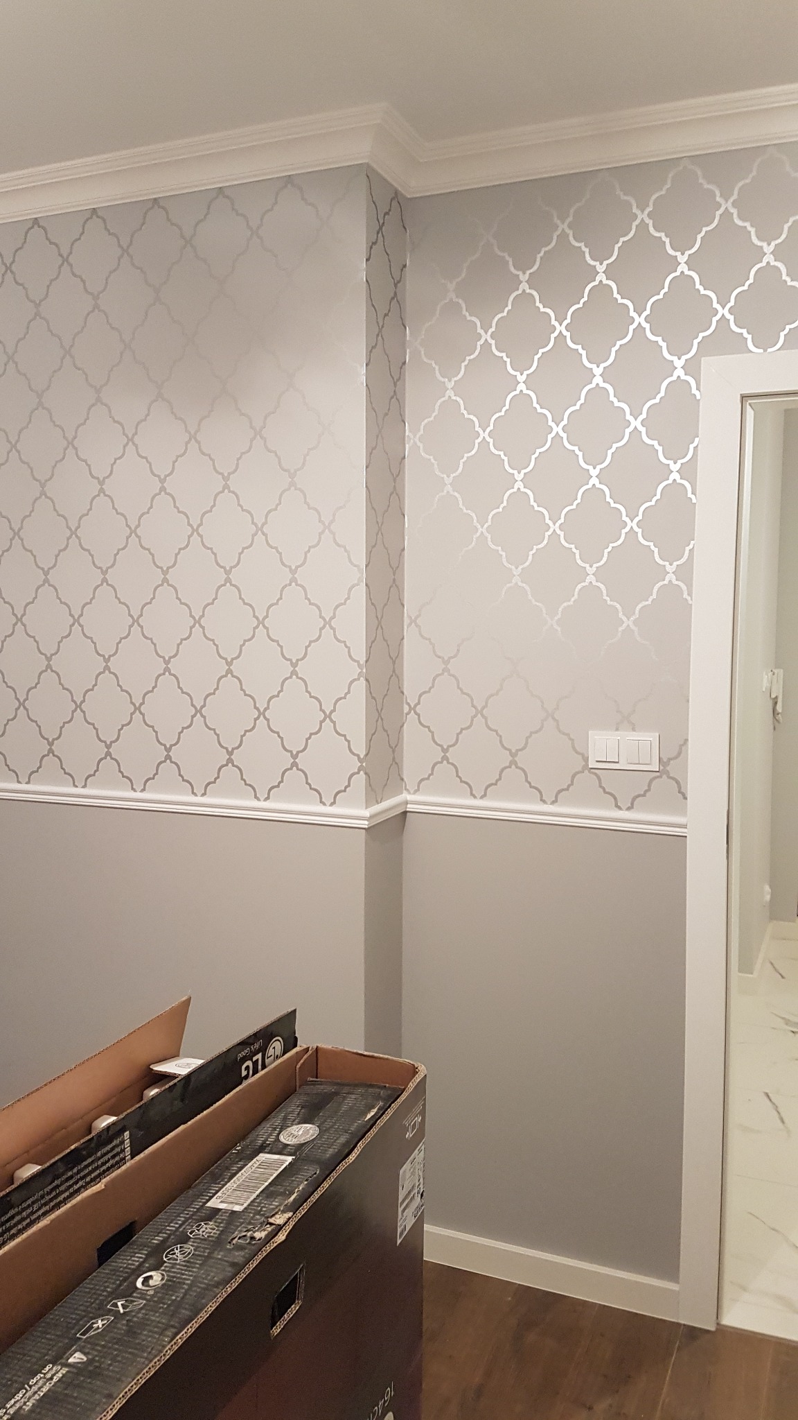 Ściana pomalowana w dolnej części na szaro, w górnej części wzór srebrnych marokańskich koniczynek, biała listwa przypodłogowa i sufitowa, kartony na podłodze, widok na marmurową podłogę...