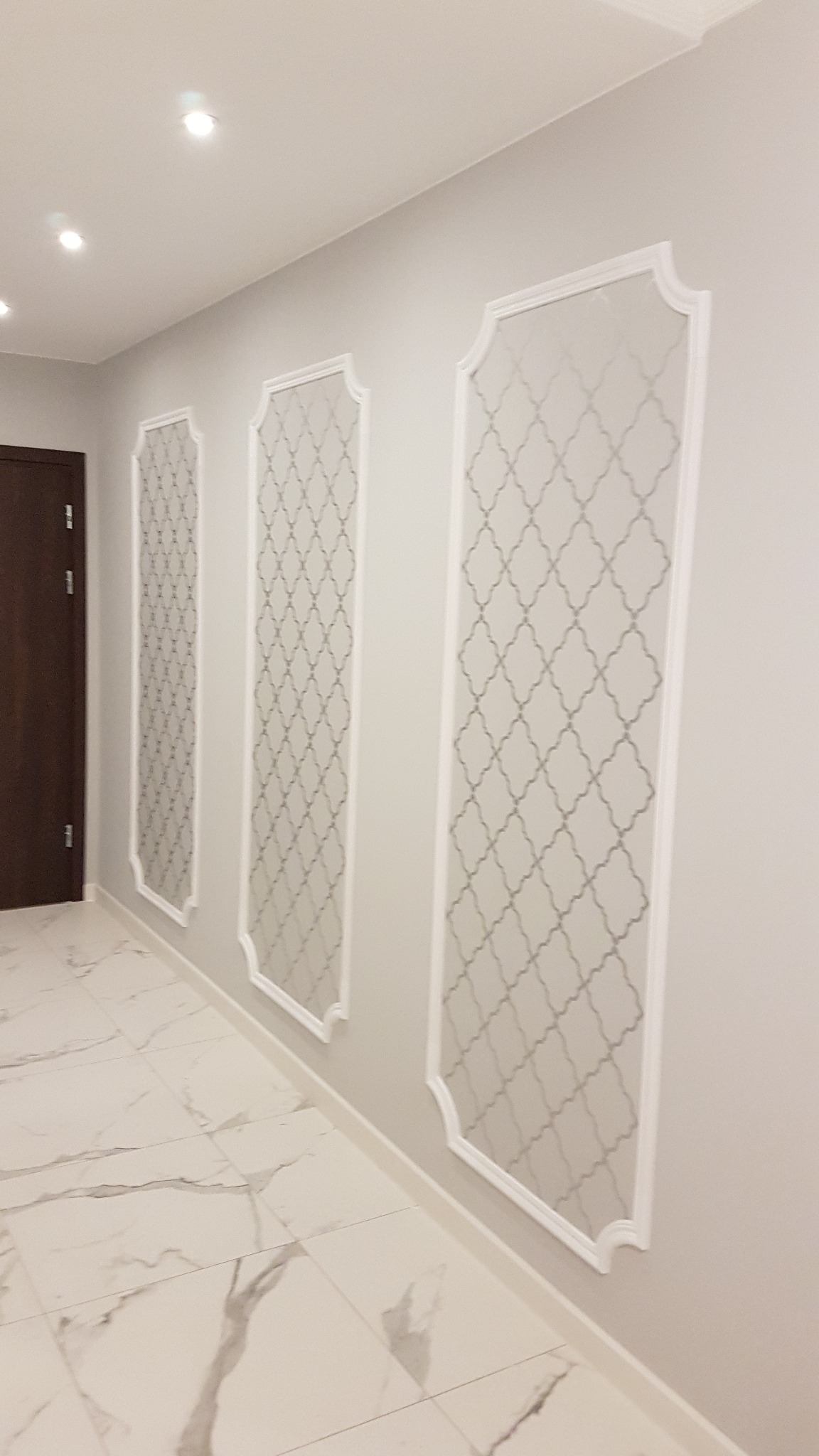 Korytarz z trzema dekoracyjnymi panelami ściennymi w białych ramach, z delikatnym geometrycznym wzorem na szarym tle, marmurowa podłoga, widoczne oświetlenie sufitowe i fragment brązowych drzwi.