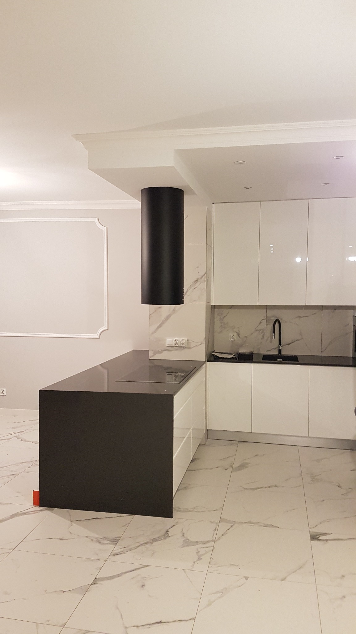 Nowoczesna kuchnia z wyspą, białe lakierowane szafki, czarny okrągły okap i marmurowe płytki podłogowe z widocznym fragmentem listwy przypodłogowej.