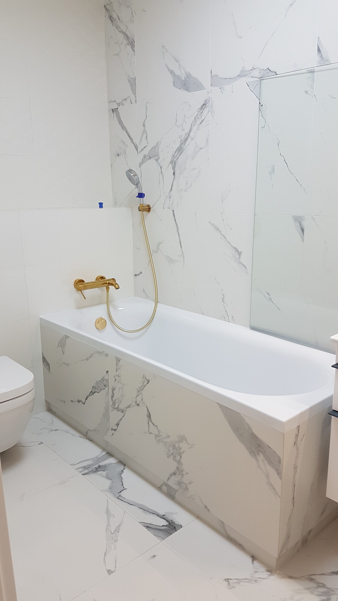 Biała wanna obudowana płytkami imitującymi marmur, złota bateria i deszczownica, ściany wyłożone białymi płytkami z szarymi żyłami, widoczny fragment toalety