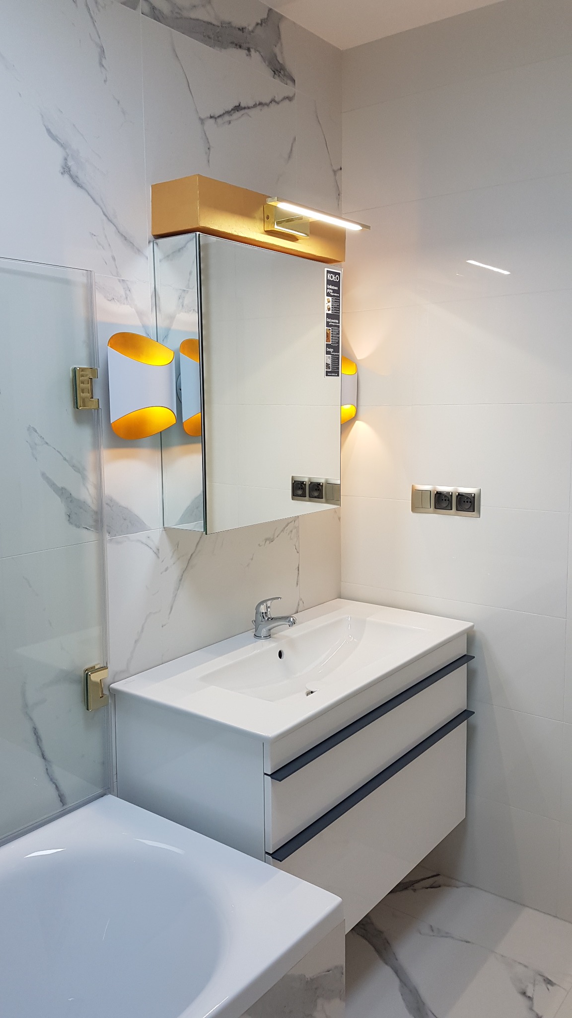 Nowoczesna łazienka z białą umywalką na szafce z szufladami, lustrem z oświetleniem LED, marmurowymi płytkami ściennymi i designerskimi lampami ściennymi.