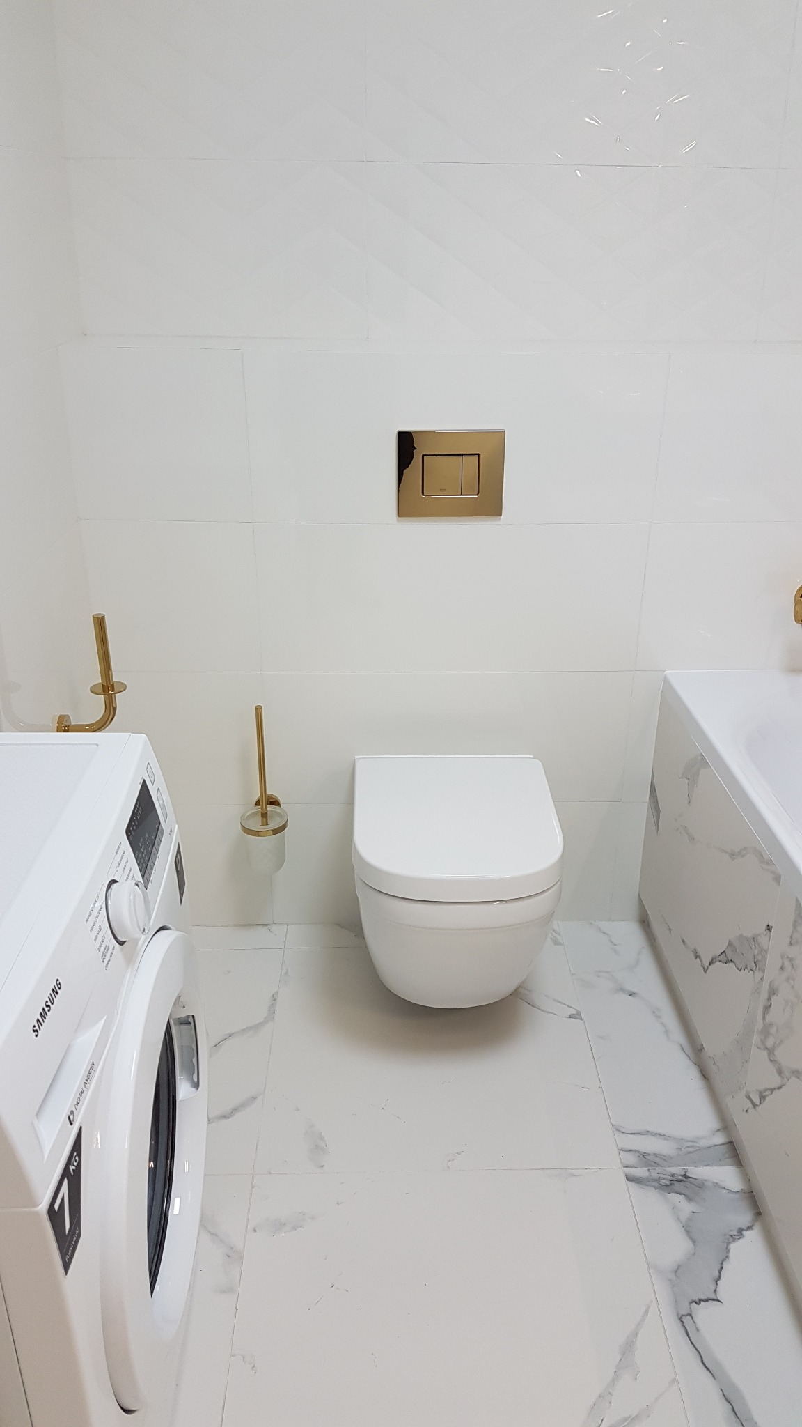 Nowoczesna łazienka z białymi płytkami i marmurową podłogą, toaleta w stylu minimalistycznym, złote akcenty w postaci przycisku spłuczki i uchwytu na papier toaletowy, pralka Samsung z boku.