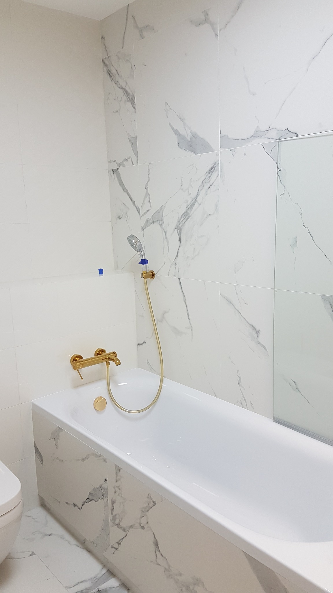 Biała wanna obudowana płytkami imitującymi marmur, złota bateria i słuchawka prysznicowa na tle ściany wyłożonej płytkami z motywem marmuru.