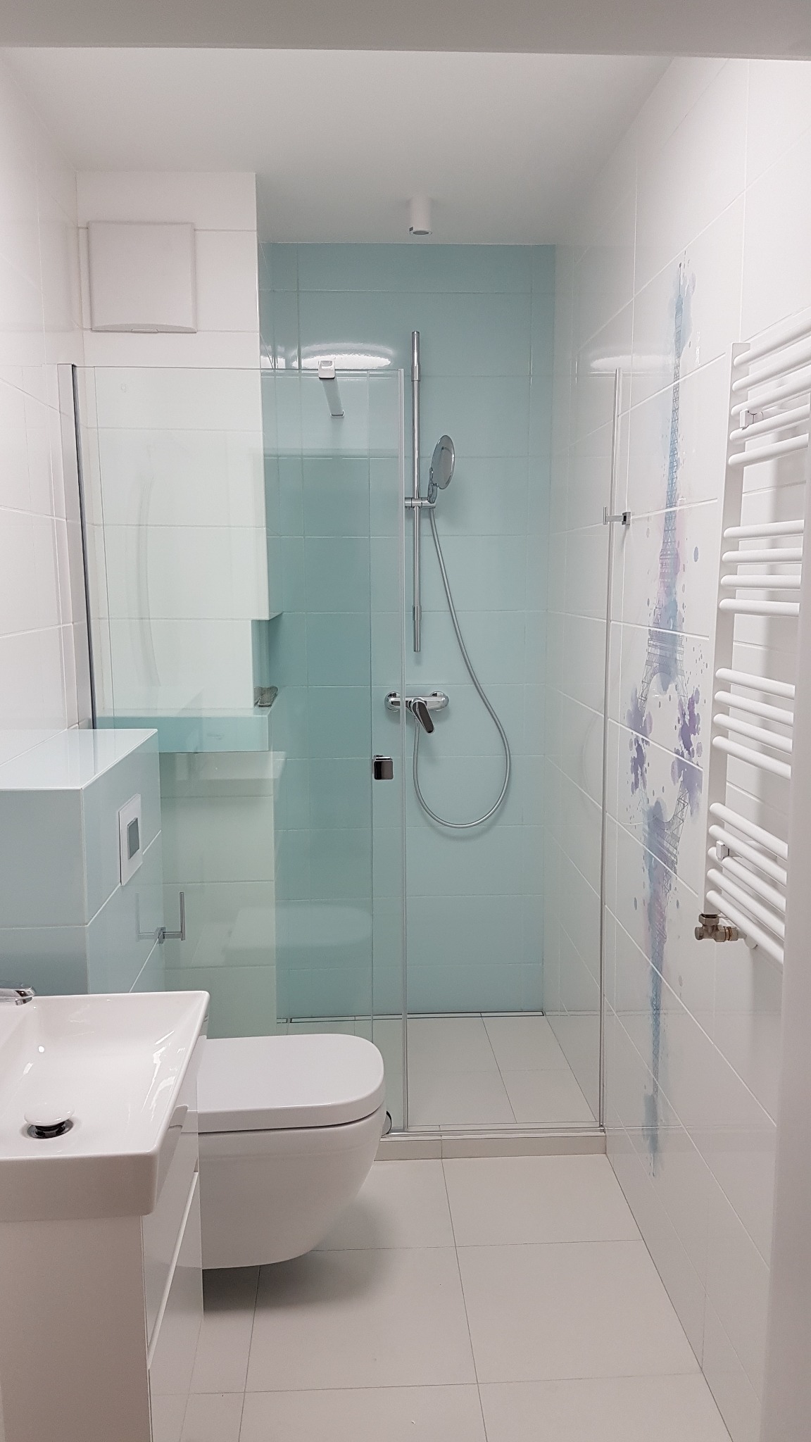 Nowoczesna łazienka z kabiną prysznicową z turkusowymi płytkami, umywalką nablatową, podwieszaną toaletą i grzejnikiem drabinkowym. Na ścianie widoczny motyw wieży Eiffla.