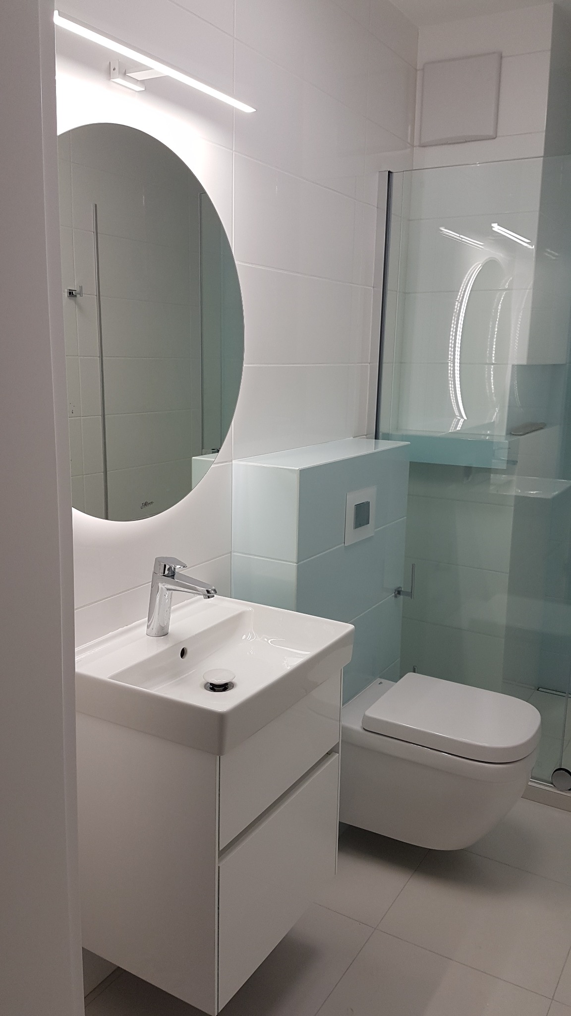 Nowoczesna, biała łazienka z okrągłym lustrem podświetlanym LED, wiszącą toaletą i kabiną prysznicową ze szklanymi drzwiami.