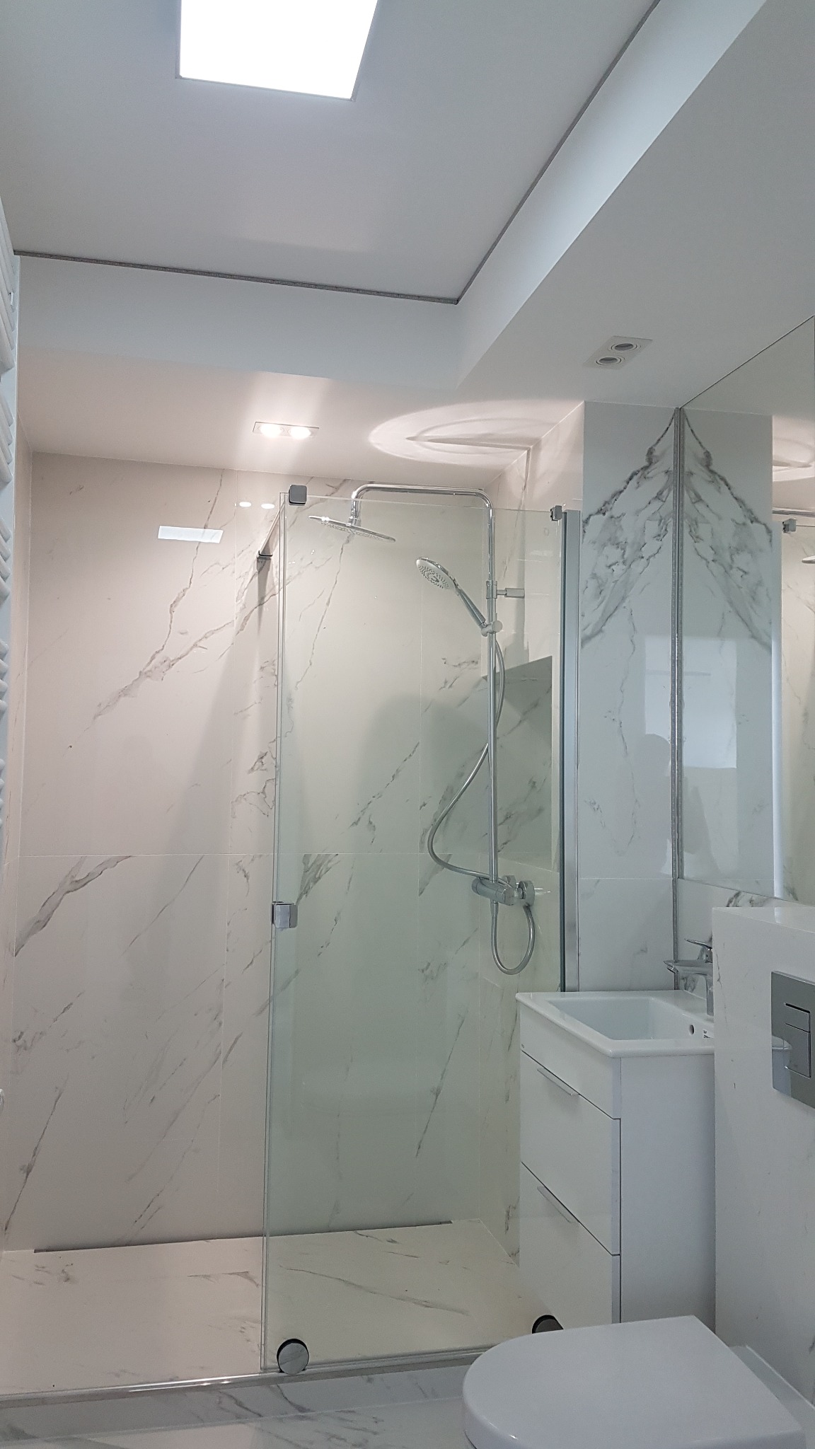 Nowoczesna łazienka z kabiną prysznicową typu walk-in, wykończona białymi płytkami imitującymi marmur, z minimalistyczną umywalką i podwieszaną toaletą.