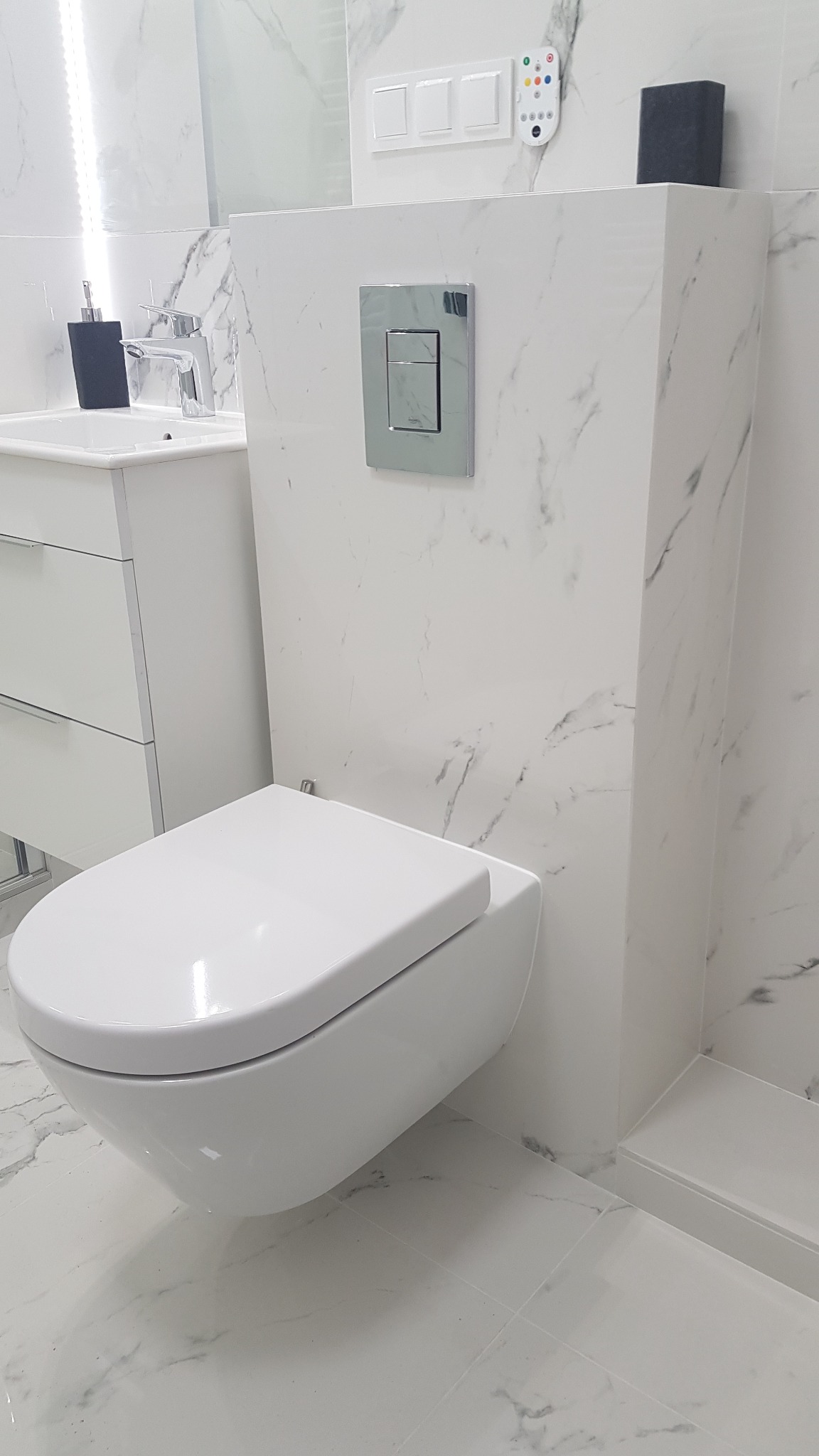 Nowoczesna, podwieszana toaleta w białej łazience wykończonej marmurowymi płytkami, z widoczną umywalką z szafką i chromowaną armaturą.