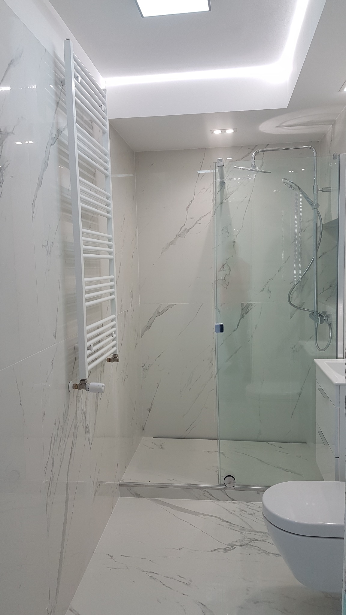 Nowoczesna łazienka wykończona białymi płytkami imitującymi marmur, z kabiną prysznicową typu walk-in, grzejnikiem drabinkowym i podświetlanym sufitem.