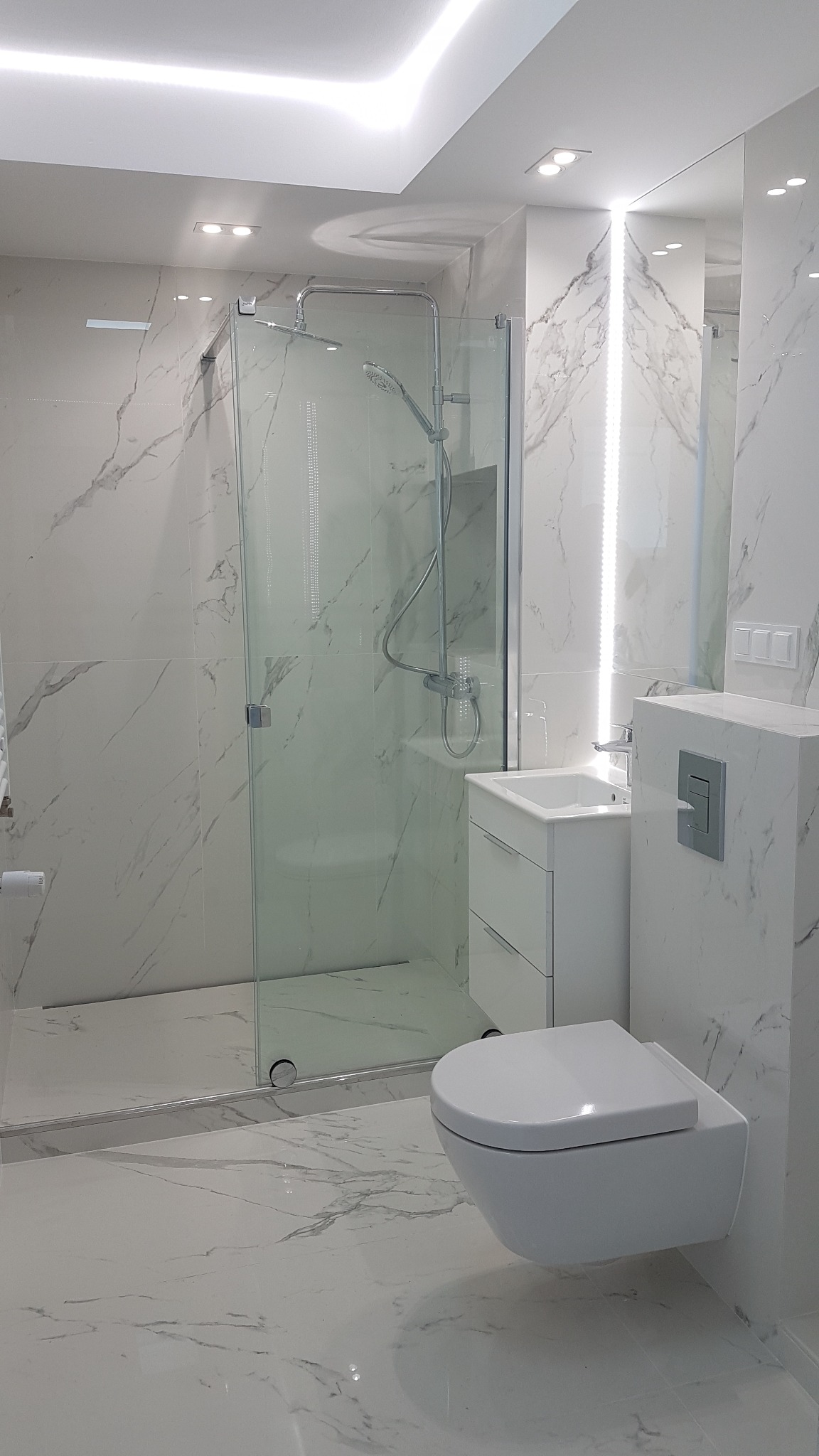 Nowoczesna łazienka wykończona białymi płytkami imitującymi marmur, z kabiną prysznicową ze szklanymi drzwiami, podwieszaną toaletą i umywalką z szafką, oświetlona listwami LED.