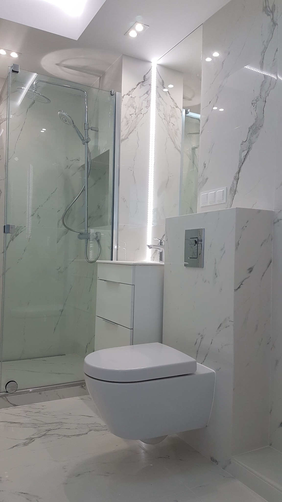 Nowoczesna łazienka wykończona białym marmurem, z podwieszaną toaletą, kabiną prysznicową ze szklanymi drzwiami i szafką z umywalką, oświetlona punktowymi lampami sufitowymi i paskiem LED.