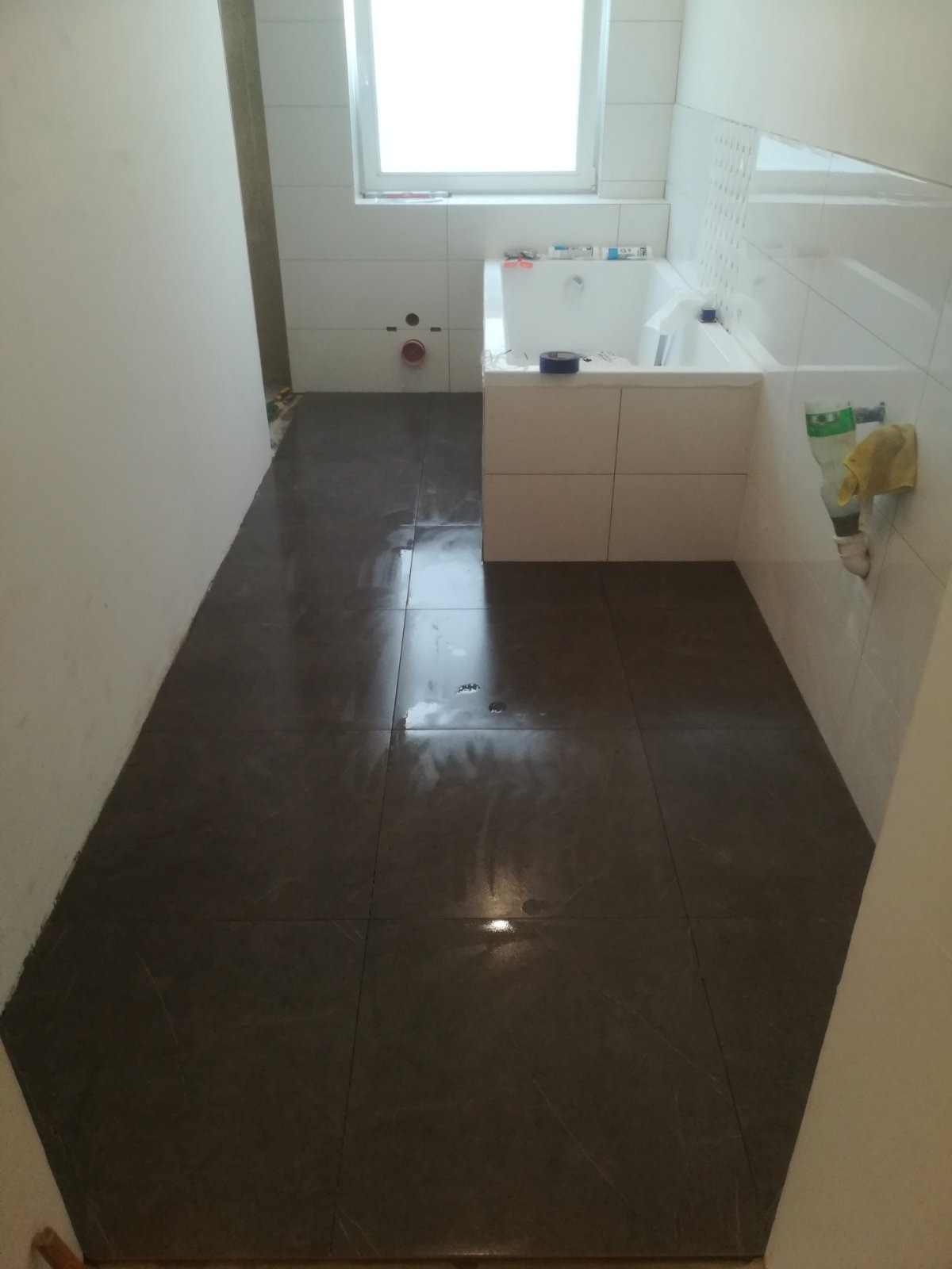 Łazienka w trakcie wykończenia: ciemne płytki podłogowe z widocznymi smugami po czyszczeniu, białe płytki ścienne, wanna obudowana płytami, okno w tle, widoczne instalacje hydrauliczne.