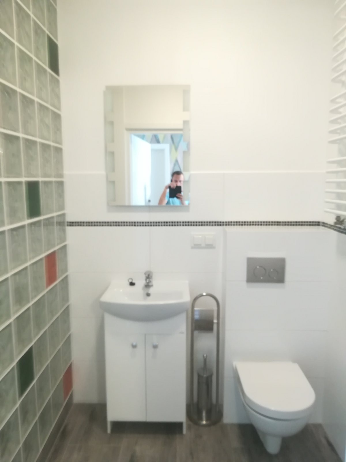 Nowoczesna łazienka z umywalką na szafce, toaletą i ścianą z luksferami, widoczne odbicie osoby robiącej zdjęcie w lustrze.