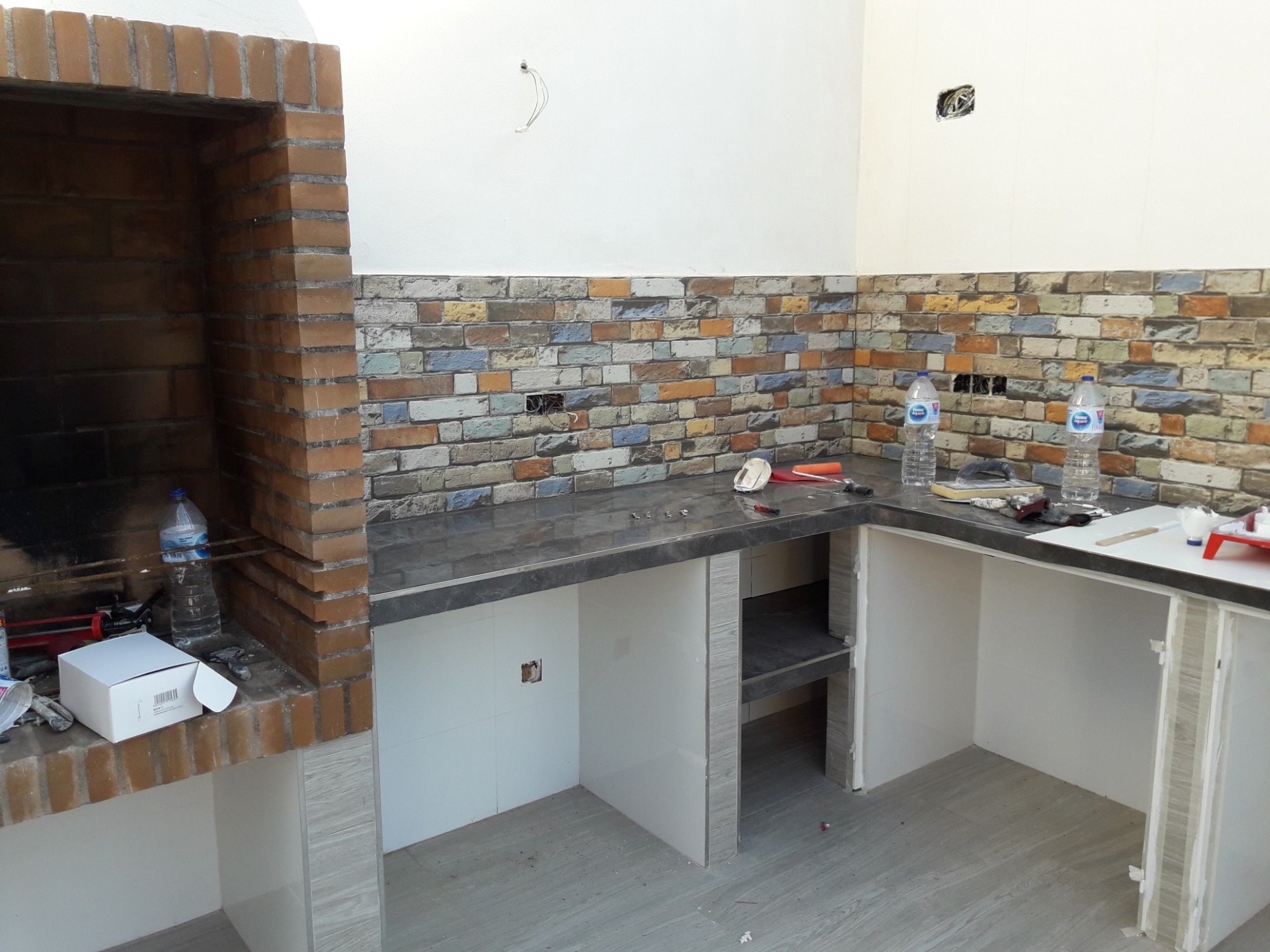 Murowany grill z cegły i fragment blatu roboczego z wzorzystą okładziną ścienną imitującą cegłę, widoczne narzędzia i materiały budowlane, wnętrze w trakcie wykańczania.