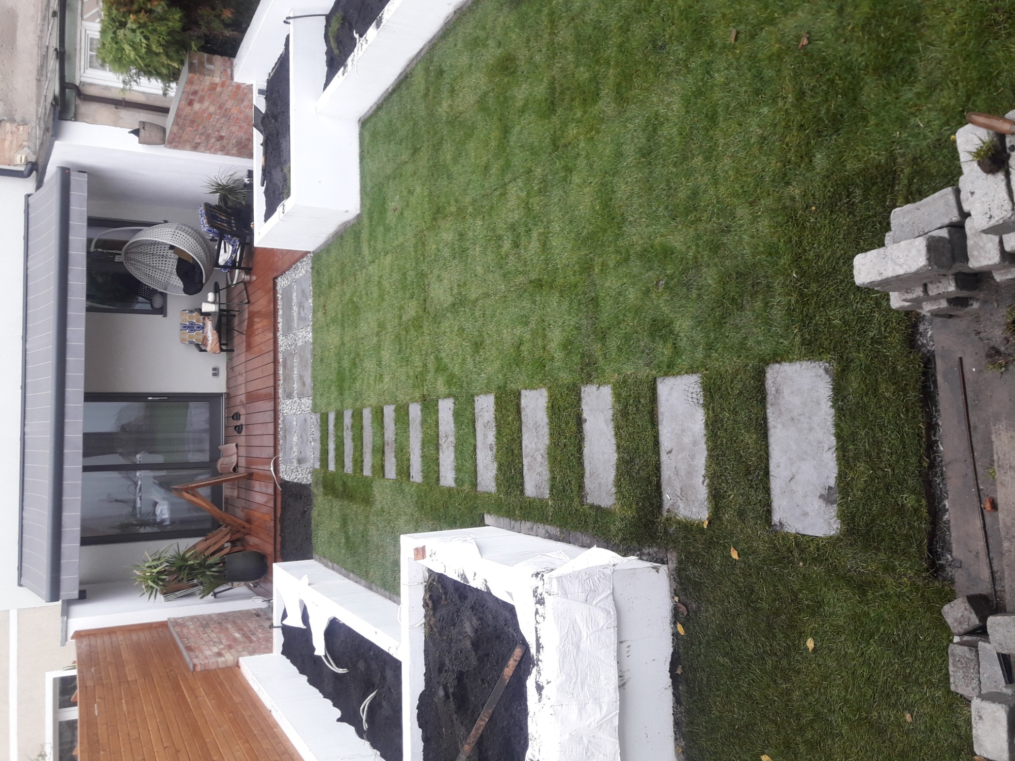 Widok z góry na ogród po remoncie z równo ułożonym trawnikiem, ścieżką z płyt betonowych i podwyższonymi rabatami kwiatowymi, w tle taras z meblami ogrodowymi.