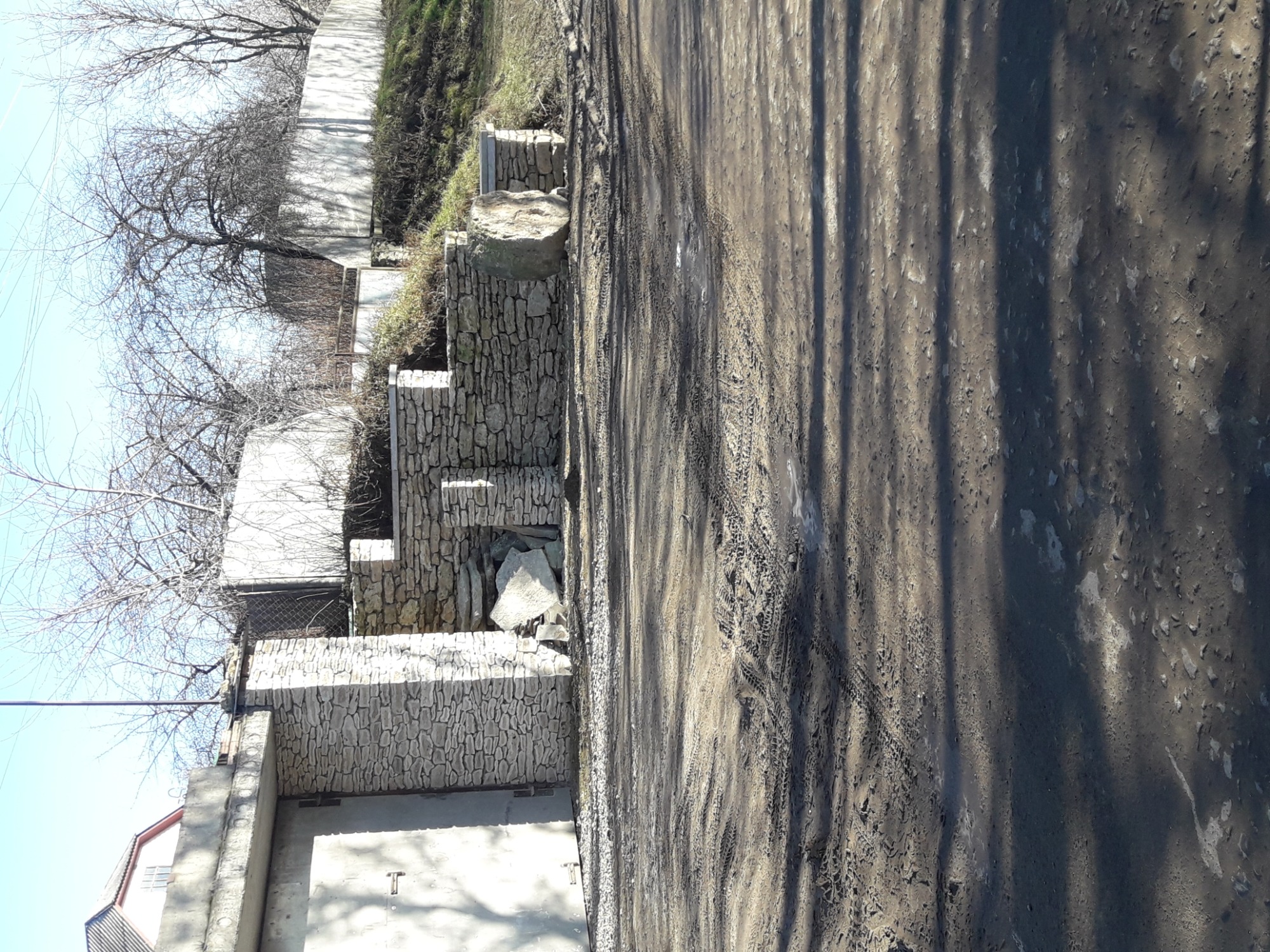 Kamienne ogrodzenie kaskadowe z elementami dekoracyjnymi, widok z boku na tle ziemistej drogi i drzew bez liści.