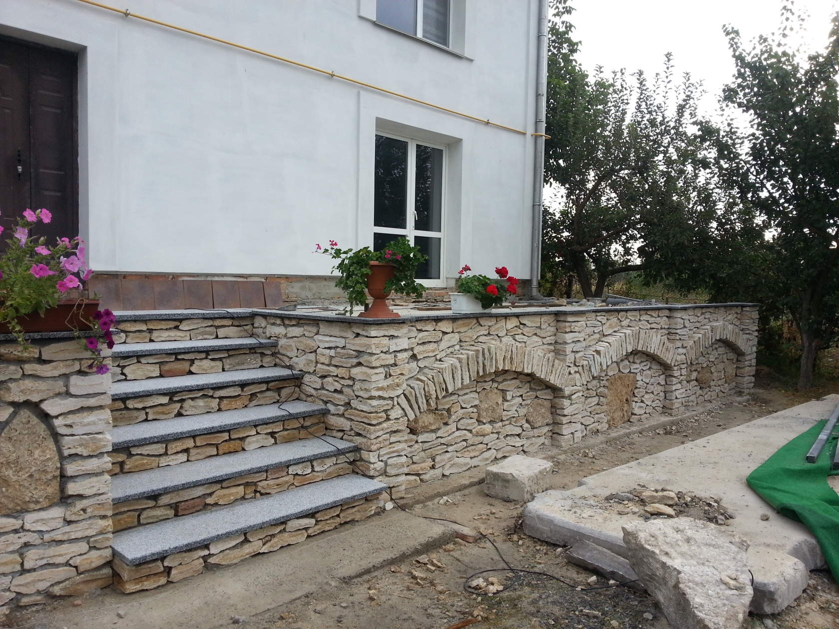 Wykończone kamienne schody wejściowe do domu z kwiatami w donicach i ozdobnym murem z kamiennymi łukami, widoczne elementy wykończeniowe na zewnątrz budynku.