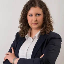 Portret kobiety w granatowej marynarce z ażurowym wykończeniem, białej koszuli i srebrnym zegarku, z założonymi rękami na tle jasnego tła.