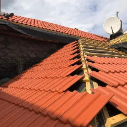 Częściowo pokryty dach z czerwonej dachówki ceramicznej, widoczne drewniane łaty i membrana dachowa, w tle komin z anteną satelitarną i fragment ściany budynku.