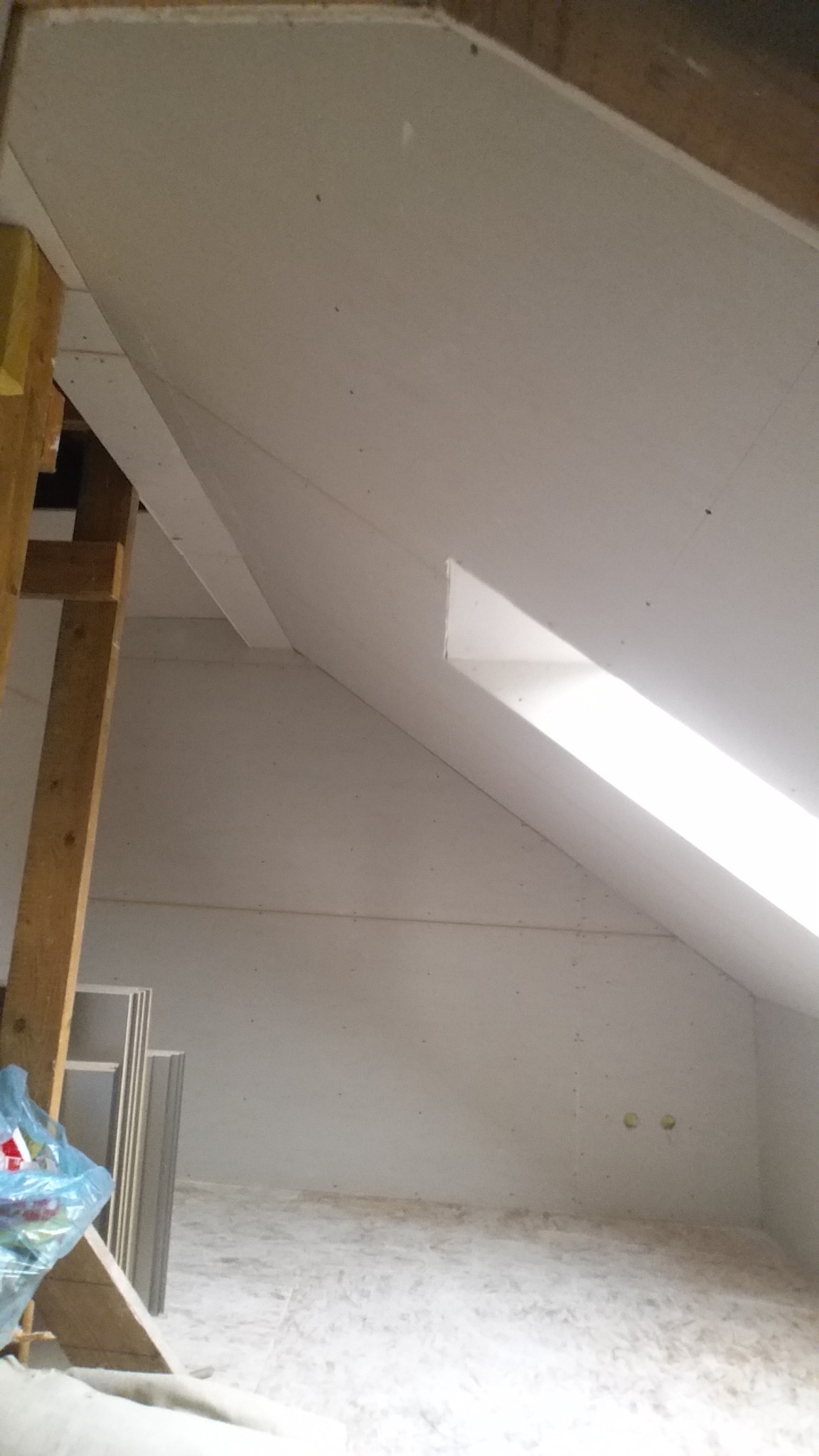 Wykończone poddasze z zabudową z płyt, widoczne drewniane elementy konstrukcyjne i fragment okna dachowego w ukośnej ścianie.