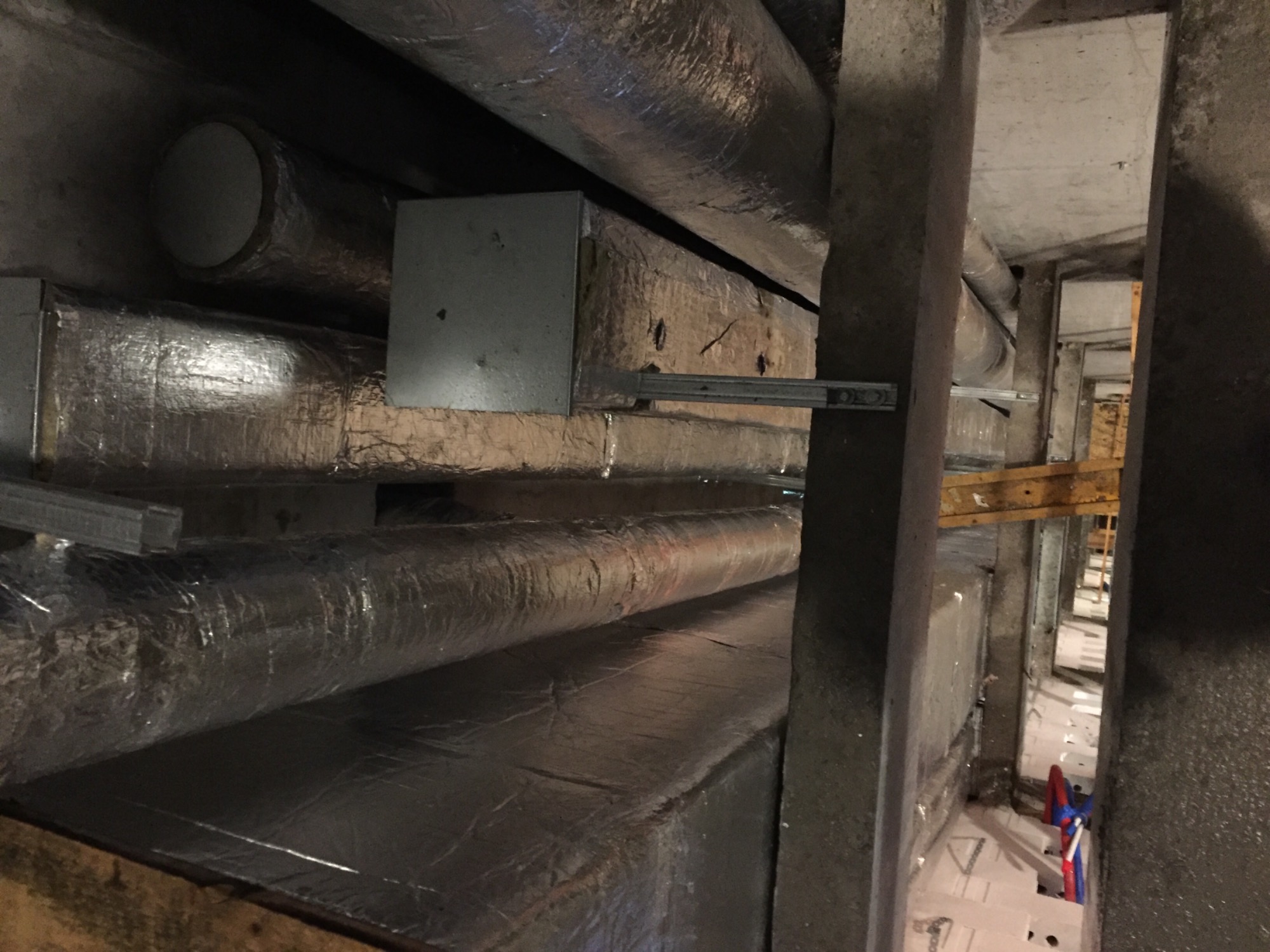 Rury wentylacyjne owinięte izolacją z folii aluminiowej, zamontowane pod sufitem w surowym pomieszczeniu technicznym, widoczne elementy konstrukcyjne i fragmenty instalacji hydraulicznej w tle.