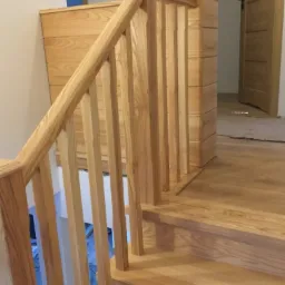 Jasne, drewniane schody z balustradą i prostymi szczeblami, widok z dołu, w tle drzwi wewnętrzne z ciemnym akcentem.