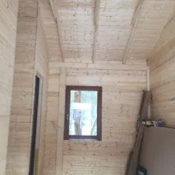 Wnętrze surowego budynku z drewnianych bali, widoczne okno z brązową ramą i przygotowane materiały budowlane przy ścianie.