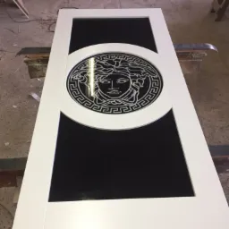 Białe drzwi z czarnym szkłem i dekoracyjnym motywem Meduzy w okrągłej ramie, leżące na warsztatowym stole.