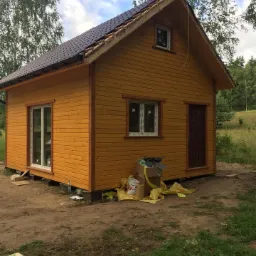Żółty drewniany budynek z brązowym dachem w trakcie budowy, widoczne elementy konstrukcyjne dachu i materiały budowlane wokół.