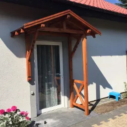 Zadaszenie nad drzwiami wejściowymi wykonane z drewna w kolorze jasnego brązu, z ozdobnymi elementami krzyżowymi, zamontowane przy białej ścianie domu z czerwonym dachem, na pierwszym planie...