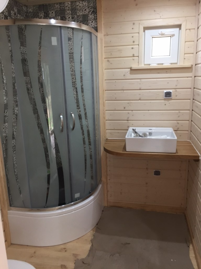 Wnętrze łazienki w domu z drewnianych bali: kabina prysznicowa z wzorzystą szybą, biała umywalka na drewnianym blacie, małe okno z białymi ramami i drewniane ściany.