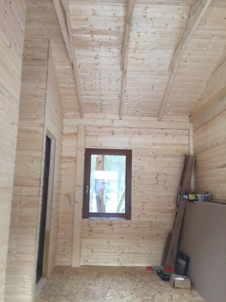 Wnętrze surowego budynku z drewnianych bali, widoczne okno z brązową ramą i przygotowane materiały budowlane przy ścianie.