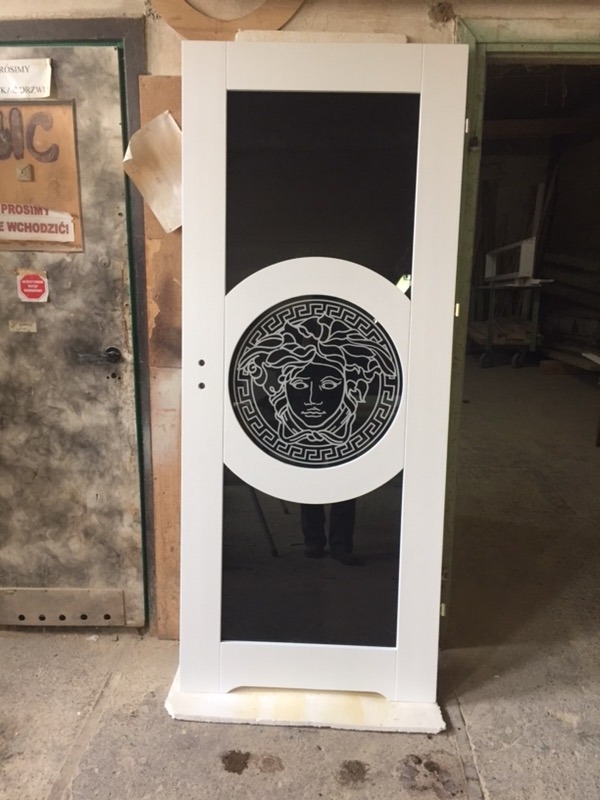 Białe drzwi wewnętrzne z czarnym szkłem i dekoracyjnym motywem Meduzy, widoczne w warsztacie stolarskim