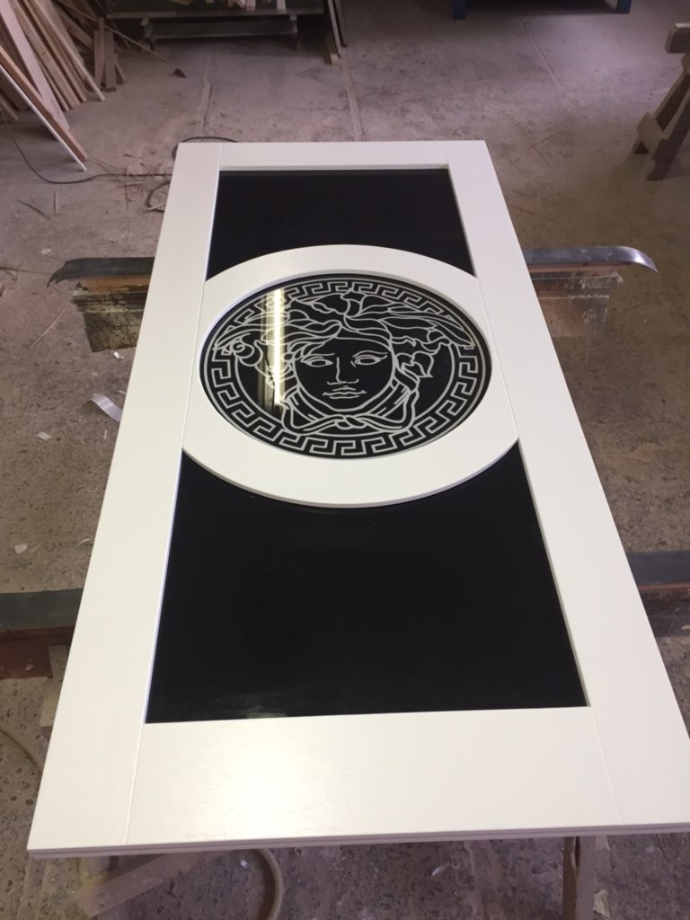 Białe drzwi z czarnym szkłem i dekoracyjnym motywem Meduzy w okrągłej ramie, leżące na warsztatowym stole.