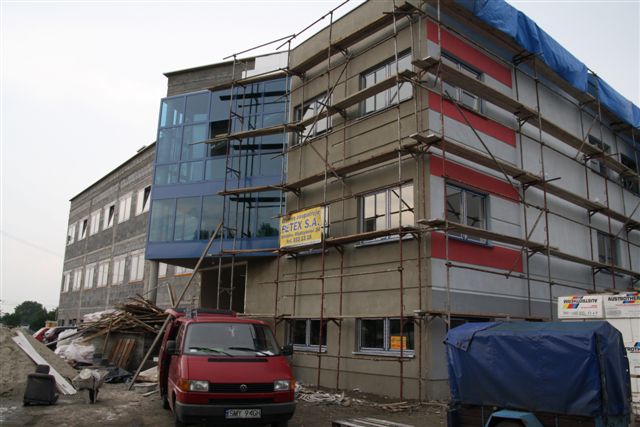 Budynek w trakcie prac wykończeniowych, elewacja częściowo pokryta tynkiem, widoczne rusztowania, czerwony bus VW Transporter na pierwszym planie.