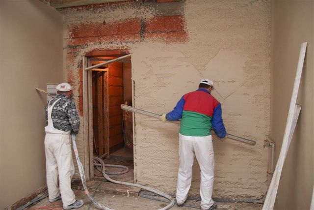 Dwóch pracowników w trakcie nakładania i wyrównywania zaprawy na ścianie wewnątrz budynku. Jeden pracownik używa agregatu, drugi wyrównuje zaprawę długą łatą. Widoczne przewody agregatu leżące...