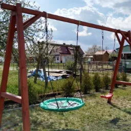 Drewniana huśtawka z okrągłym siedziskiem w kolorze zielonym i pojedynczym czerwonym siedziskiem na tle ogrodu z domami i drzewami.