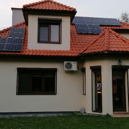 MAWBUD plus Marek Wojtasik - Dom jednorodzinny z czerwoną dachówką, na której zamontowano panele fotowoltaiczne; na ścianie budynku widoczna jednostka zewnętrzna klimatyzacji.