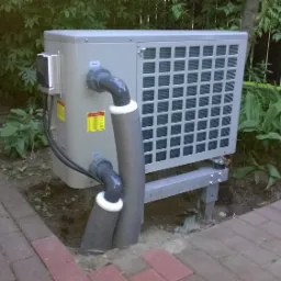 Zewnętrzna jednostka pompy ciepła zamontowana na metalowej konstrukcji, z widocznymi rurami w izolacji, umieszczona na tle zieleni i kostki brukowej.