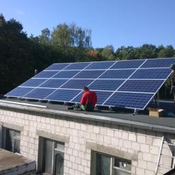 MAWBUD plus Marek Wojtasik - Montaż paneli słonecznych na dachu budynku z białej cegły. Jeden pracownik siedzi na dachu, drugi stoi obok paneli, obok karton, drabina oparta o ścianę.