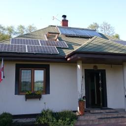 MAWBUD plus Marek Wojtasik - Dom jednorodzinny z panelami słonecznymi i kolektorami słonecznymi na dachu pokrytym zieloną dachówką, widoczne okno dachowe, flaga Polski, kwiaty w oknach i donicach.