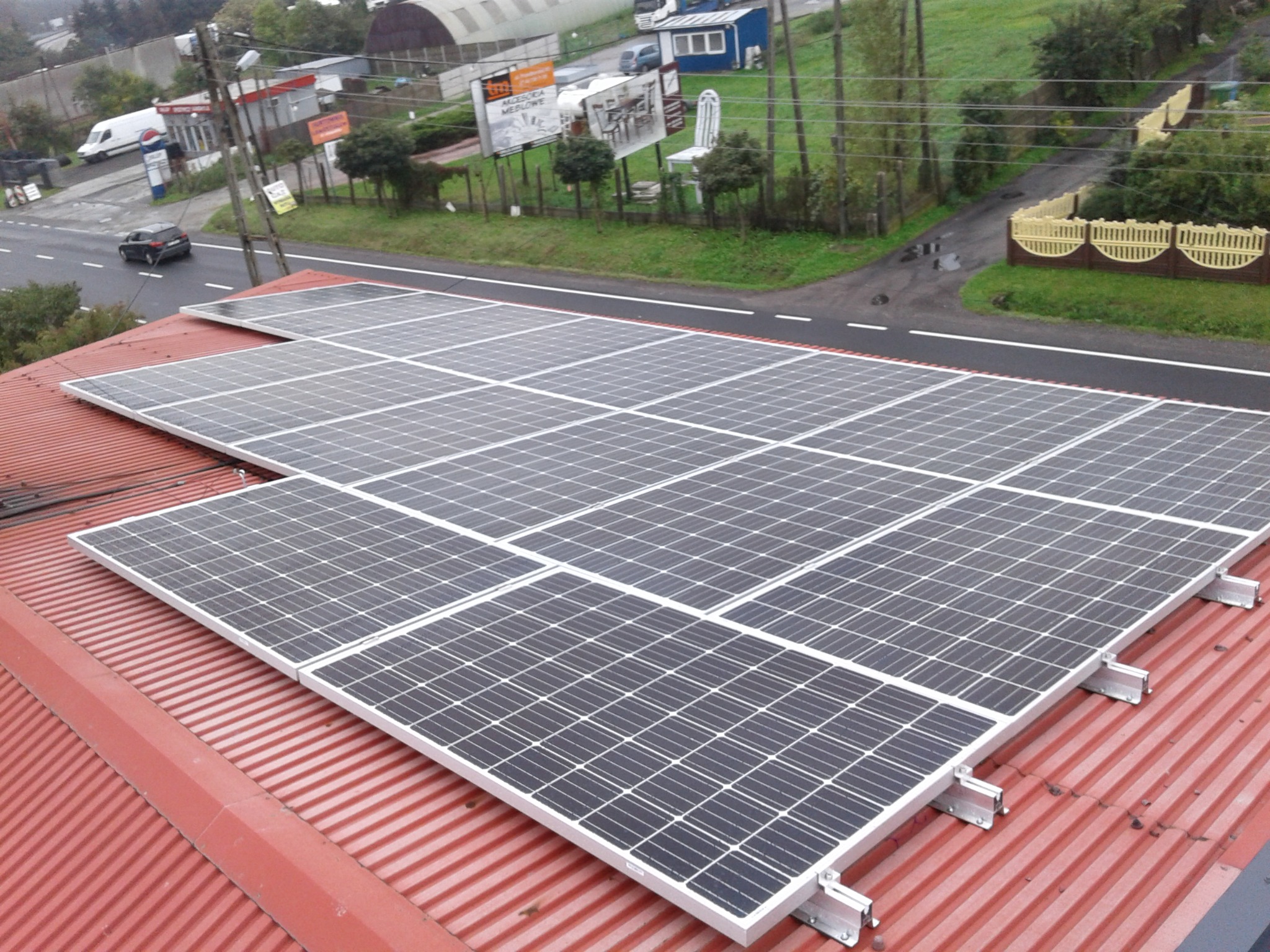 Instalacja fotowoltaiczna na czerwonym, blaszany dachu budynku, widok z góry, z ulicą i billboardami w tle.