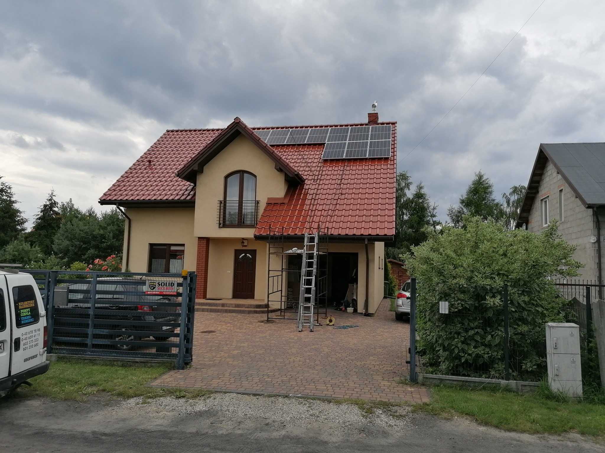 Dom jednorodzinny z czerwoną dachówką, na której zamontowane są panele słoneczne, widoczne rusztowanie i drabina, sugerujące trwający montaż instalacji.