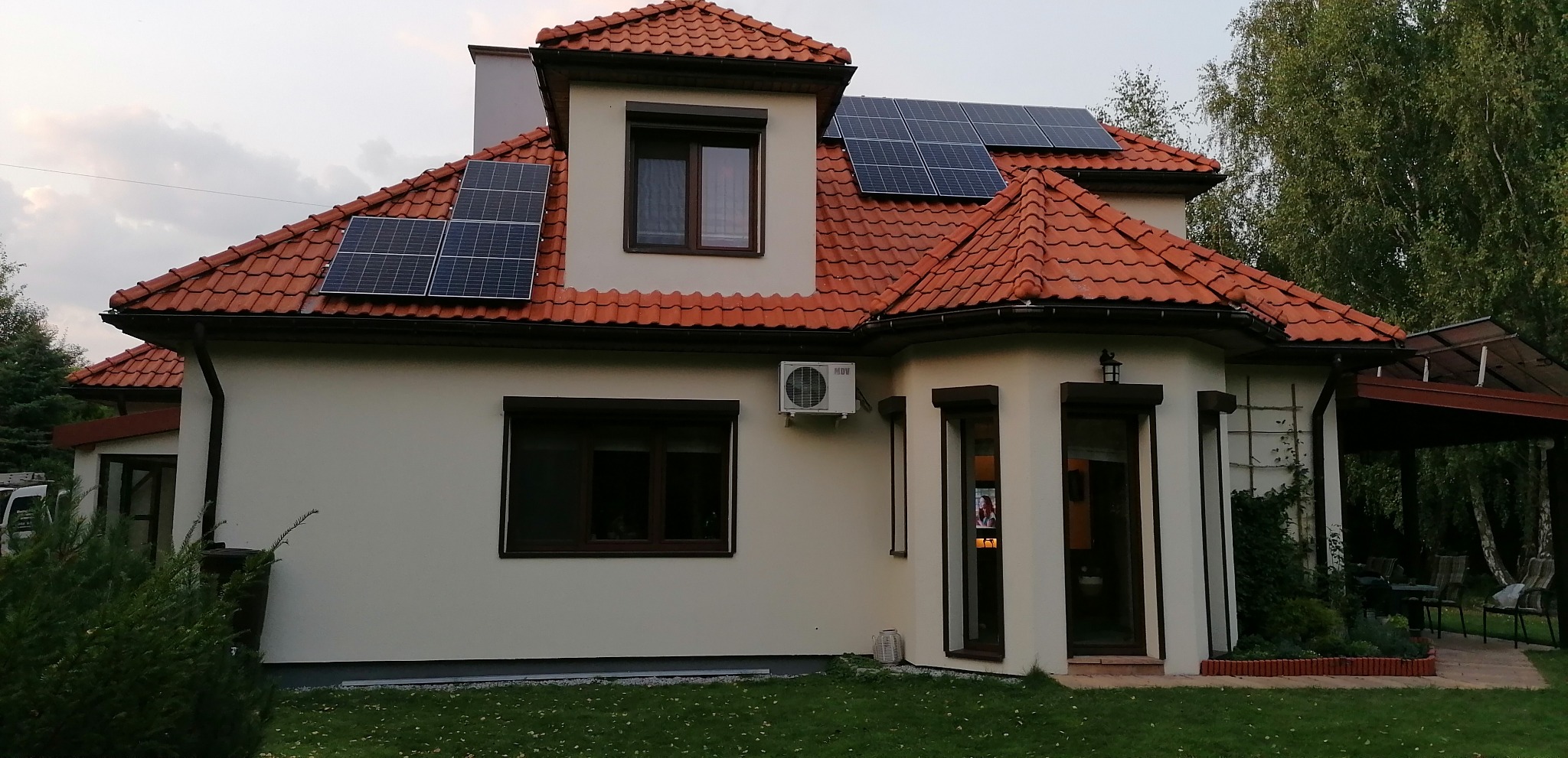 Dom jednorodzinny z czerwoną dachówką, na której zamontowano panele fotowoltaiczne; na ścianie budynku widoczna jednostka zewnętrzna klimatyzacji.