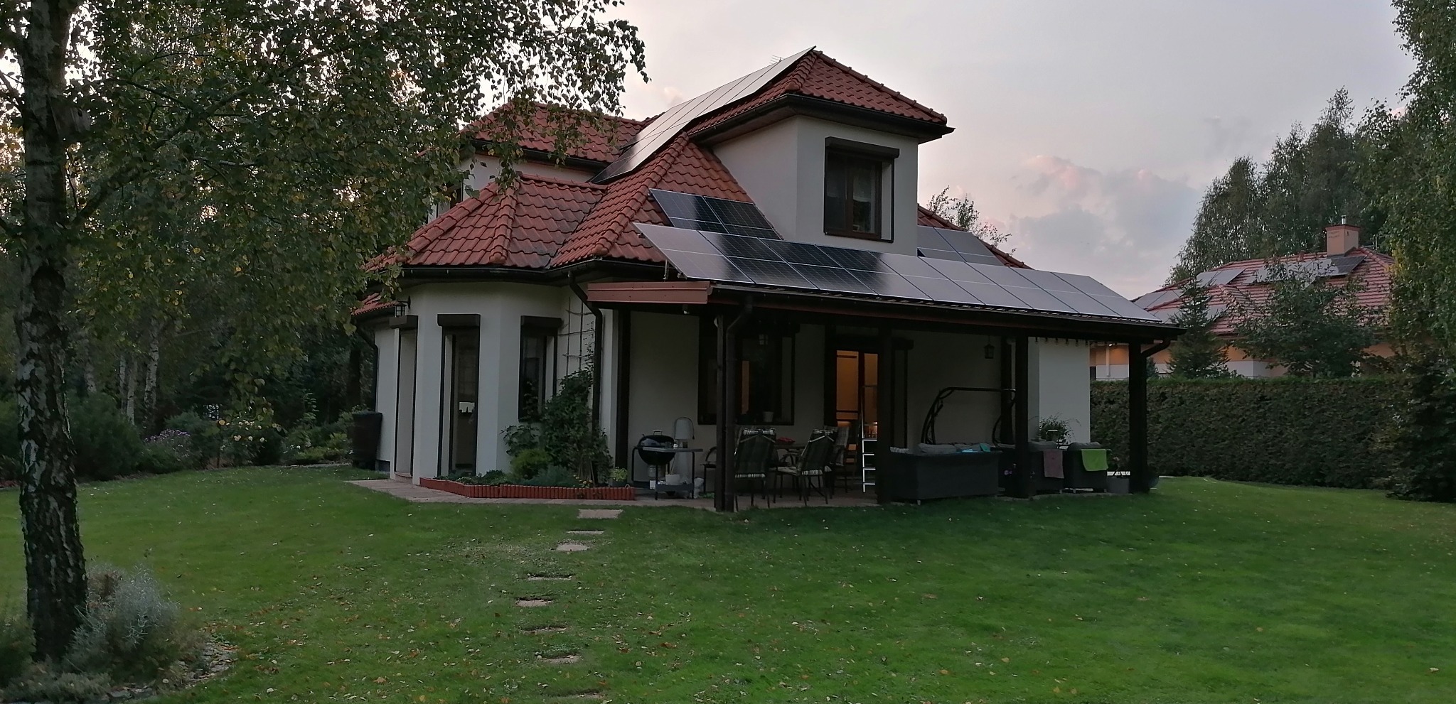 Dom jednorodzinny z panelami słonecznymi na dachu i zadaszeniu tarasu, widok z ogrodu w lekko pochmurny dzień.