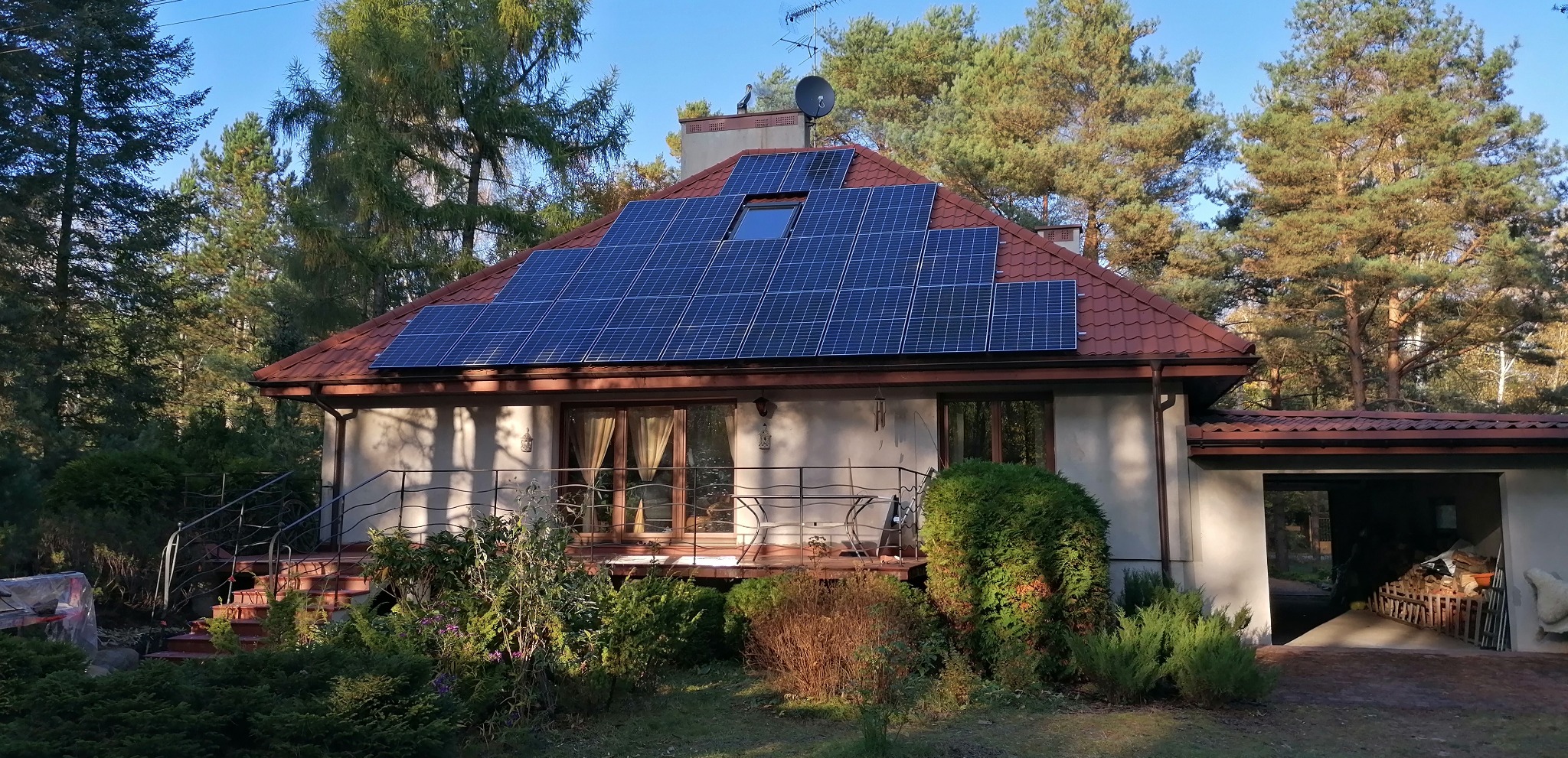 Dom jednorodzinny z panelami słonecznymi na dachu, otoczony zielenią i drzewami, z widocznym fragmentem garażu po prawej stronie.