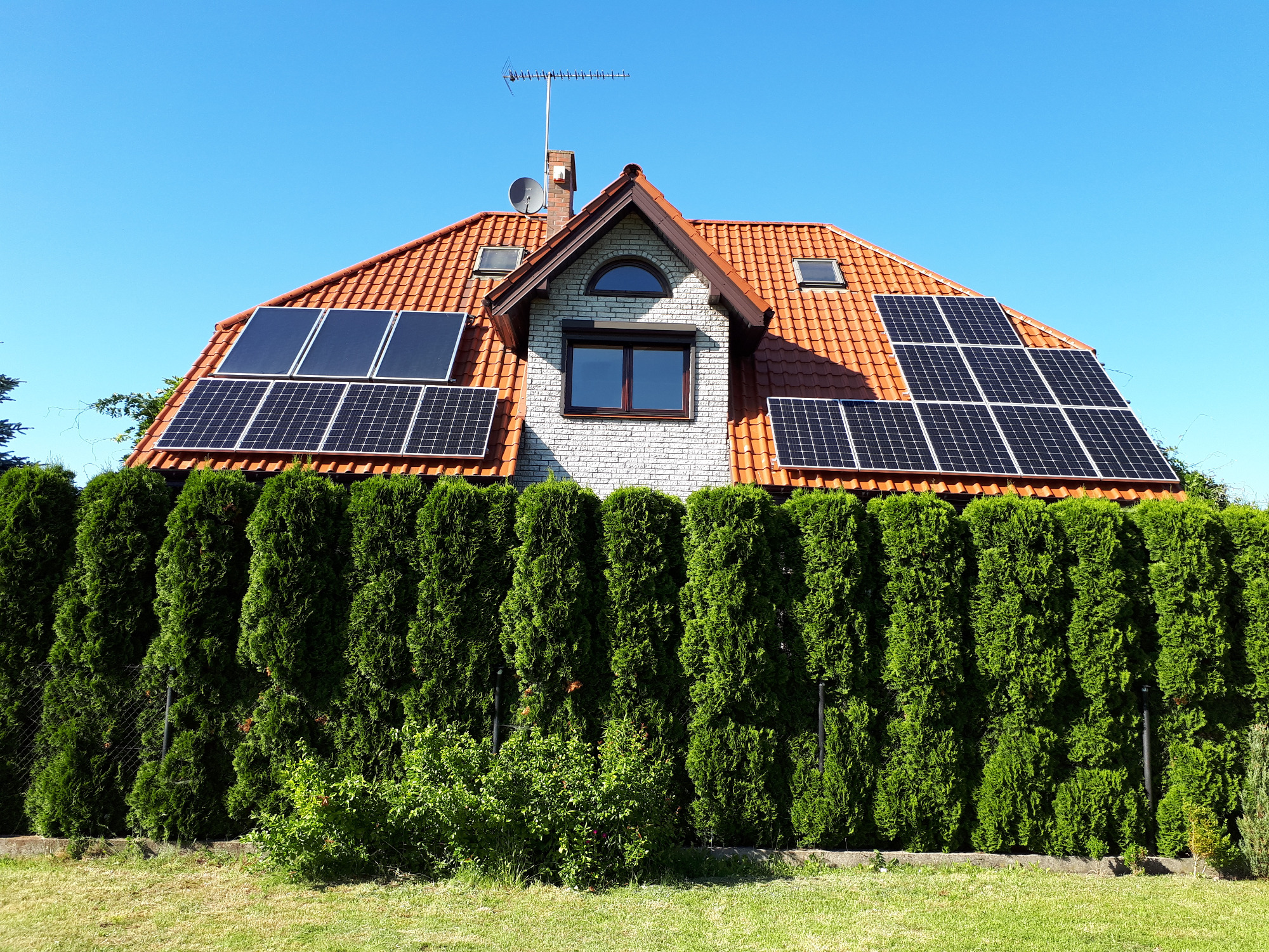 Dom jednorodzinny z czerwoną dachówką i zamontowanymi panelami fotowoltaicznymi, widoczny zza wysokiego żywopłotu.