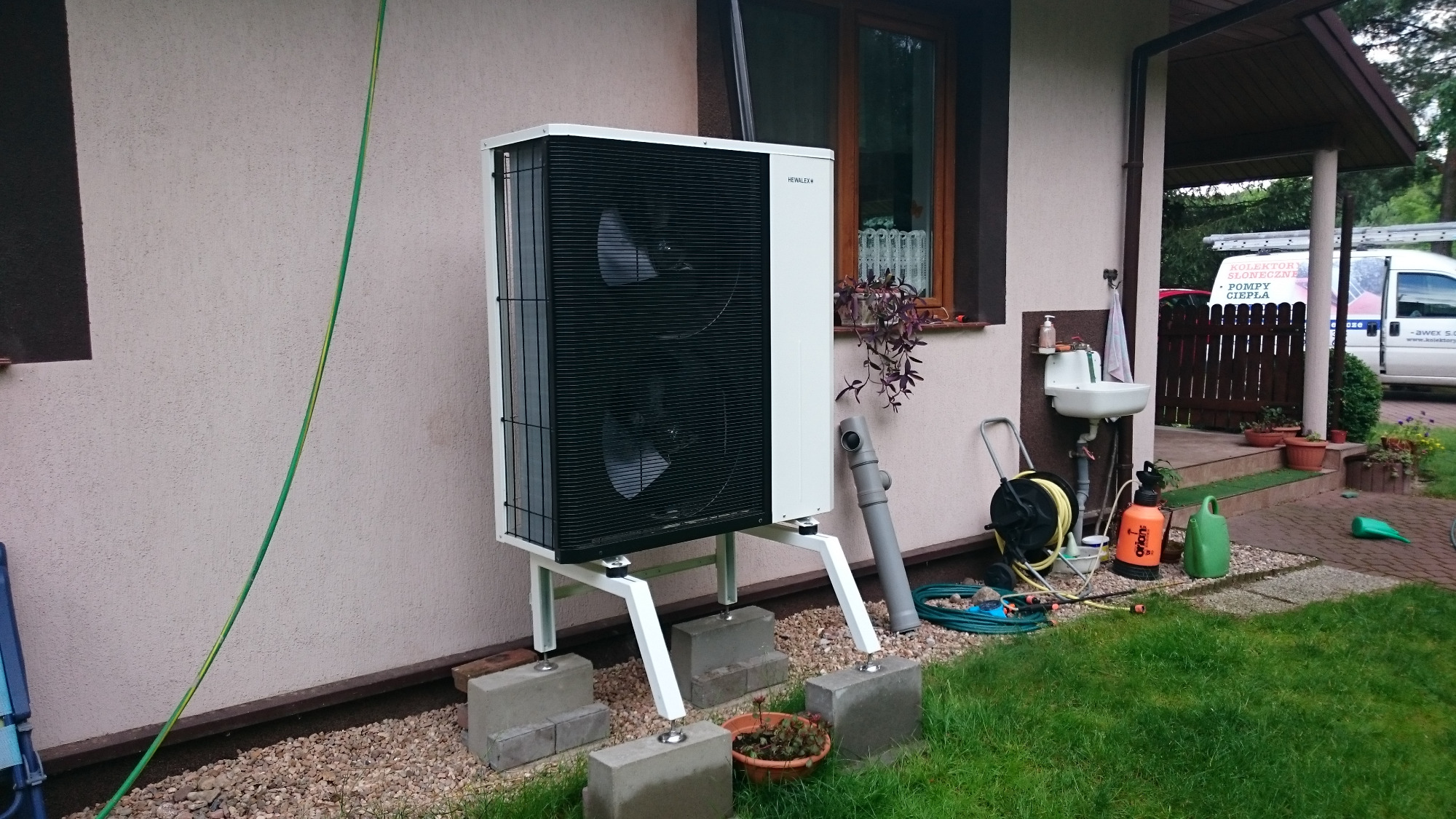 Biała pompa ciepła powietrze-woda marki Hewalex zamontowana na betonowych bloczkach przed budynkiem z beżową elewacją, widoczne akcesoria montażowe i ogród w tle.