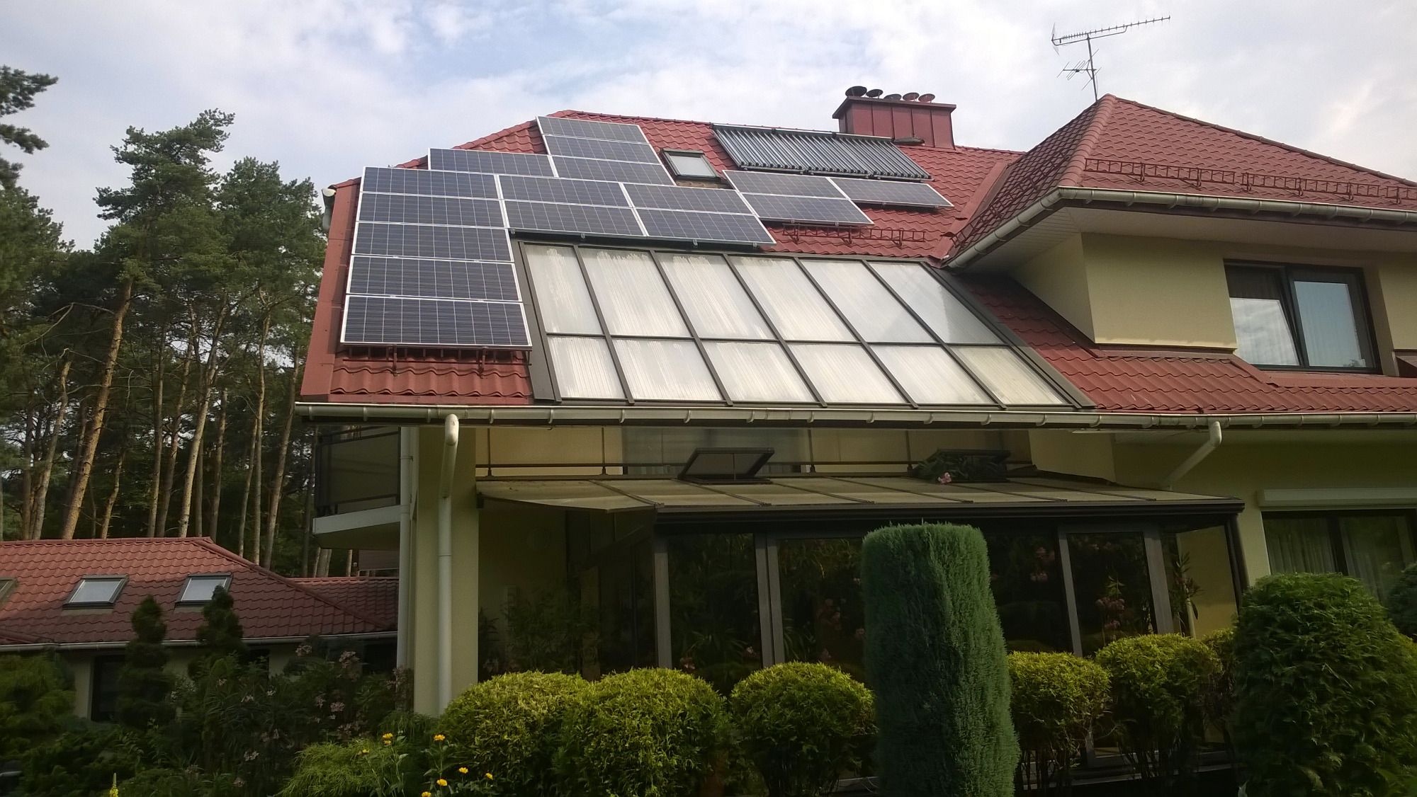 Dom jednorodzinny z panelami słonecznymi na dachu z czerwonej dachówki, dodatkowo kolektory słoneczne i przeszklona weranda.