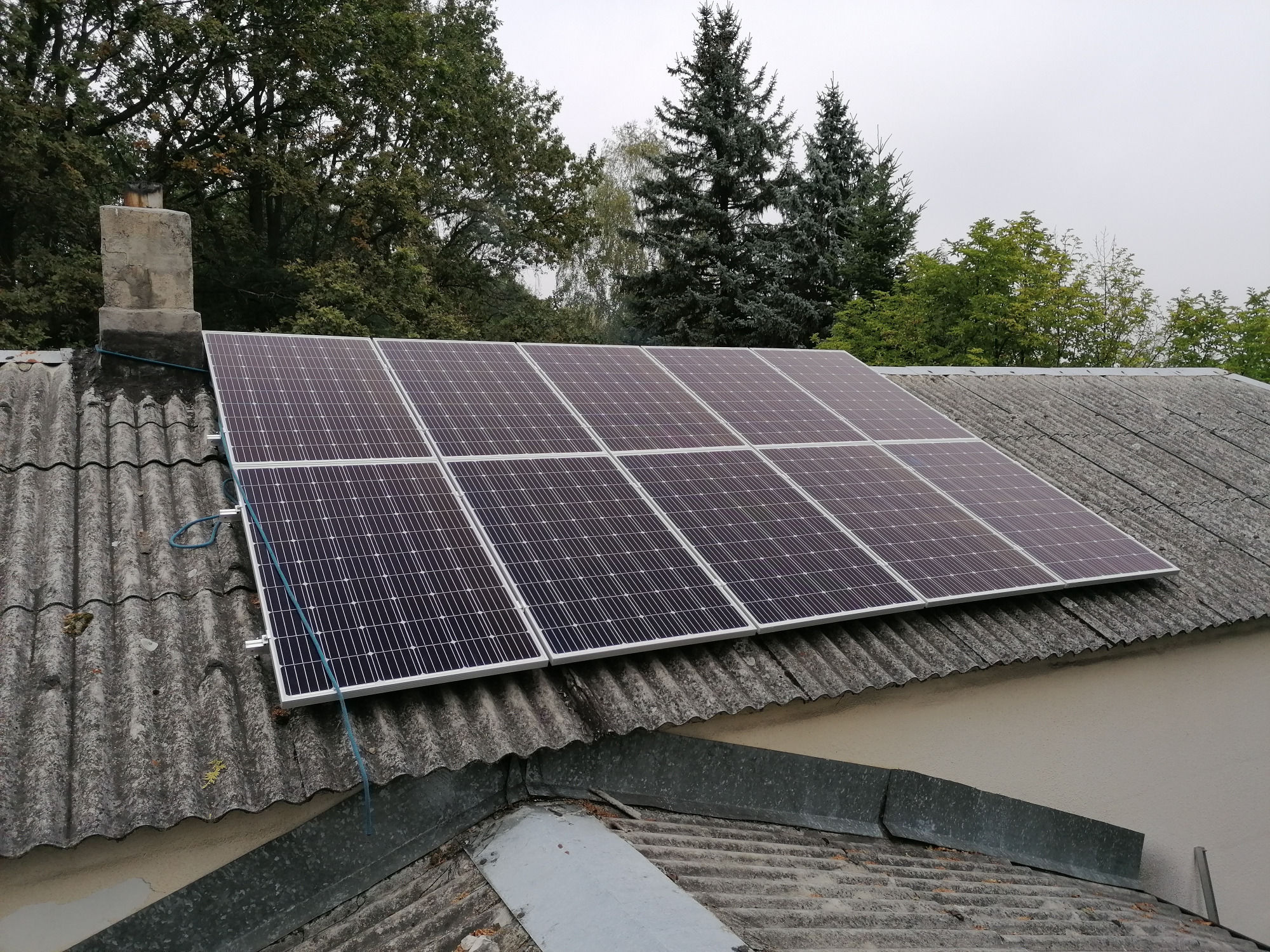 Panele słoneczne zamontowane na dachu pokrytym eternitem, widoczny komin i fragment orynnowania. W tle drzewa.